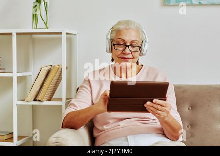 Porträt einer modernen älteren Frau, die ein digitales Tablet mit Kopfhörern verwendet, während sie auf dem Sofa in einer minimalistischen Inneneinrichtung sitzt Stockfoto