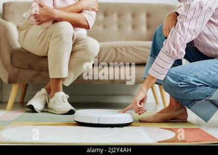 Ausgeschnittene Aufnahme einer jungen Frau, die den Roboter-Staubsauger andrückt, während sie der älteren Verwandten Smart-Home-Technologie zeigt, um Platz zu kopieren Stockfoto