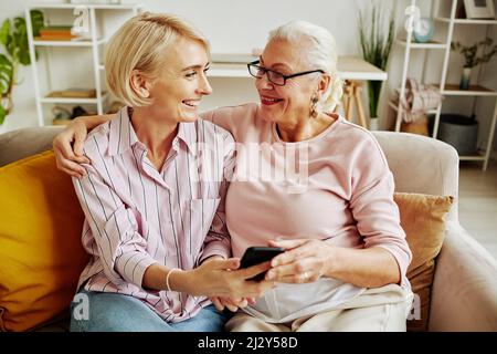 Porträt einer lächelnden jungen Frau, die die ältere Mutter anschaut, während sie in einer gemütlichen Szene auf der Couch sitzt Stockfoto