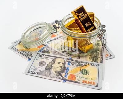 Eine Nahaufnahme von 100-Dollar-Scheinen und ein Glas Goldbarren auf einer weißen Oberfläche Stockfoto