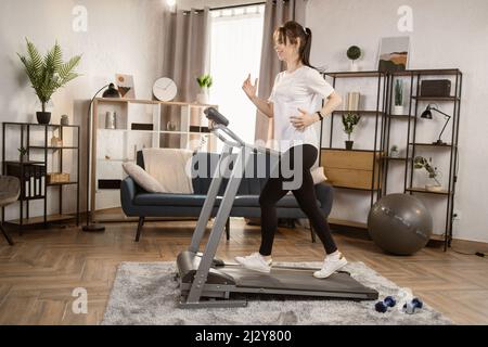 Attraktive junge kaukasische Sportlerin, die auf dem Laufband läuft. Tun Fitness-Übung. Sportliche und muskulöse Frau trainieren aktiv in der modernen Wohnung. Sport Frau Training zu Hause. Seitenansicht Stockfoto