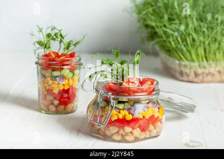 Gesunder hausgemachter Salat im Glas. Vegetarischer Salat mit Kichererbsen und Gemüse, Erbsensprossen in Gläsern auf weißem Holzhintergrund Stockfoto