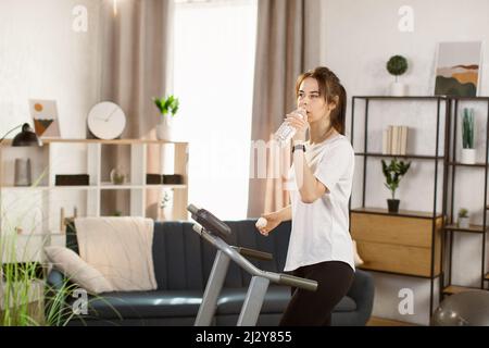 Fit athletische Frau läuft auf dem Laufband, während Wasser aus einer Flasche trinken und ihre Fitness-Übung. Muskulöse Frau trainiert zu Hause aktiv. Sport Mädchen Training im Fitness-Club. Seitenansicht Stockfoto