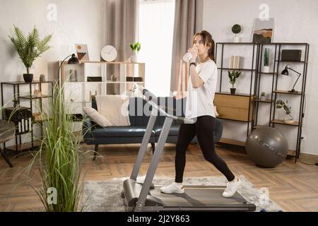 Fit athletische Frau läuft auf dem Laufband, während Wasser aus einer Flasche trinken und ihre Fitness-Übung. Muskulöse Frau trainiert zu Hause aktiv. Sport Mädchen Training im Fitness-Club. Seitenansicht Stockfoto