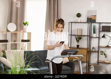 Junge fit Frau macht Übungen auf dem Laufband. Schöne junge Sportlerin mit einem Smartphone, das im Fitnessstudio auf dem Laufband läuft. Verbessern Sie Ihr Training. Stockfoto