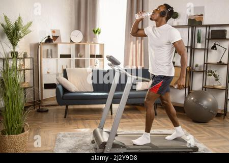 Fröhlicher junger afroamerikanischer Athlet mit Kopfhörern, der in der modernen Wohnung während des intensiven Workouts auf dem Laufband in Flaschen Wasser trinkt und dabei Lieblingslied genießt. Stockfoto