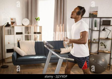 Glücklicher junger afroamerikanischer Athlet mit Kopfhörern, der während intensiver Arbeit Flaschenwasser trinkt und dabei Lieblingslied beim Laufen auf dem Laufband genießt Stockfoto