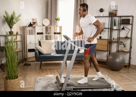 Fröhlicher afrikanischer Mann mit modernen Kopfhörern, der zu Hause auf einem Laufband läuft. Männlicher Läufer, der Cardio-Training gesund macht Lebensstil. Sportlich schlanker Mann Schwitzen Training in der Wohnung während der Freizeitbeschäftigung Stockfoto