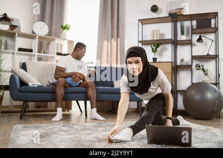 Eine arabische Frau im Kopftuch auf dem Kopf macht im Wohnzimmer zu Hause Gymnastik, während ein afrikanischer Mann auf dem Sofa sitzt und ein multinationaler kleiner Sohn in den Armen sitzt. Genießen Sie Gesunden Lebensstil, Copy Space Stockfoto