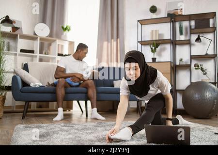 Eine arabische Frau im Kopftuch auf dem Kopf macht im Wohnzimmer zu Hause Gymnastik, während ein afrikanischer Mann auf dem Sofa sitzt und ein multinationaler kleiner Sohn in den Armen sitzt. Genießen Sie Gesunden Lebensstil, Copy Space Stockfoto