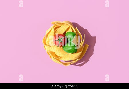 Draufsicht mit einem Nudelnest auf violettem Hintergrund in hellem Licht. Rohe Pappardelle mit Kirschtomate und Basilikumblättern. Stockfoto