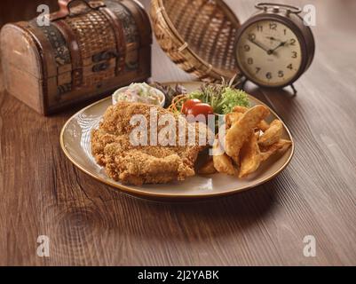 Knuspriges Hühnchen-Kotelette mit Pommes in einem Gericht, das auf einem dunklen Holztisch isoliert ist. Seitenansicht der Speisen in singapur Stockfoto