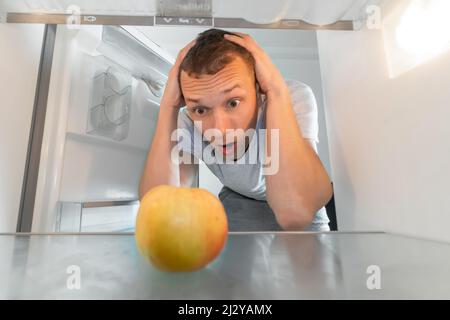 Trauriger Mann hält seinen Kopf und schaut auf den einzigen Apfel im Kühlschrank, die Kamera ist drin. Konzept von Hunger, Dients. Stockfoto