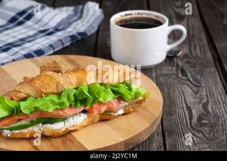 Frühstückssandwich, Croissant mit rotem Fisch, Lachs, Quark, Gurken und Salat auf dunklem Holzhintergrund mit Kaffee und Serviette. Stockfoto