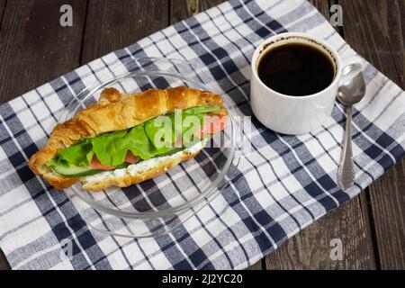 Frühstückssandwich, Croissant mit rotem Fisch, Lachs, Quark, Gurken und Salat auf dunklem Holzhintergrund mit Kaffee und Serviette, Blick von einem Stockfoto