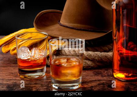 Das Wild-West-Konzept mit Cowboy-Hut, Seil-Lasso, Lederhandschuhen, zwei Gläsern Whiskey auf den Felsen und einer Flasche Bourbon auf einem Holztisch in Stockfoto