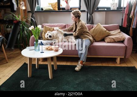 Mädchen mit Armprothese streicheln ihren Hund auf dem Sofa während der Freizeit im Wohnzimmer Stockfoto