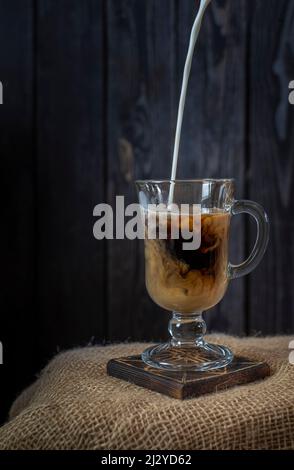 Milch wird in rustikalem Stil mit Kaffee auf dunklem Holzboden in ein Glas gegossen Stockfoto