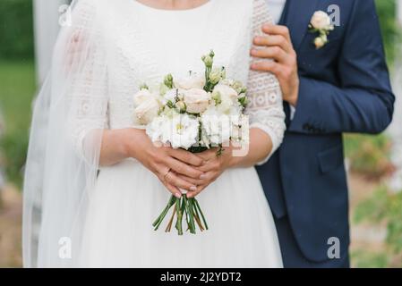 Die Braut hält in ihren Händen einen schönen Strauß aus zarten Blumen und grünen Blättern. Das Brautpaar umarmt, die Hände der Braut und des Bräutigams Stockfoto