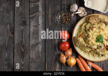 Usbekischer Pilaf mit Ente. Reis mit Stücken Entenfleisch mit Kochzutaten, Zwiebeln, Karotten, Knoblauch, Paprika und Tomaten. Flach auf dunklem Holz legen Stockfoto