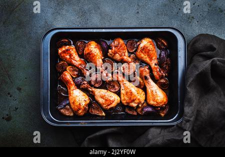 Gebackene Hähnchenbeine, Drumsticks mit süßen Feigen und Zwiebeln in balsamischer Marinade auf schwarzem Backblech, rostiger grüner Tischhintergrund, Draufsicht Stockfoto