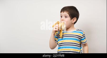 Ein 4-jähriger Junge isst eine Banane. Langes Banner mit Platz für Text auf weißem Hintergrund. Früchte und Vitamine für Kinder im Vorschulalter Stockfoto