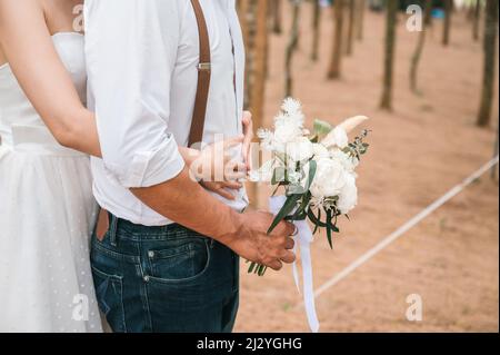 Nahaufnahme der Brücke und des Bräutigams, der vor der Hochzeit im Wald steht und umarmt, mit Blumenstrauß. Hochzeitskleid, fröhliche Paar Stockfoto