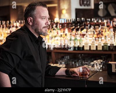 Porträt eines hübschen reifen Mannes, der ein Glas ordentlichen Whiskys in der Bar oder im Pub hält. Stockfoto