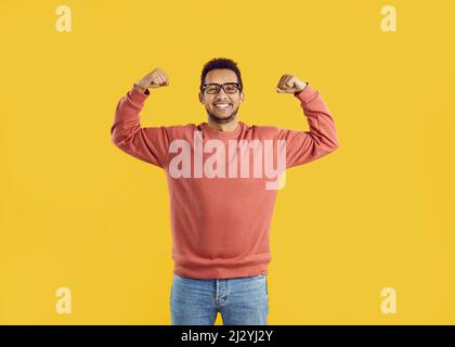 Fröhlicher, lustiger junger Mann mit Sweatshirt und Brille, der seine Arme beugte, um zu zeigen, wie stark er ist Stockfoto