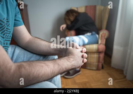 Mann in bedrohlicher Haltung und Frau in einem Sessel zusammengeruckt weinend, Gewalt gegen Frauen. Stockfoto