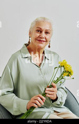 Minimalporträt einer lächelnden älteren Frau, die Blumen hält und die Kamera anschaut, während sie im Studio posiert Stockfoto