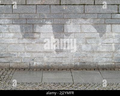 Nasse graue Steinwand aus rechteckigen Blöcken. Der gepflasterte Bürgersteig befindet sich vor einer Fassade. Das Äußere des Gebäudes ist ein abstrakter Hintergrund. Stockfoto