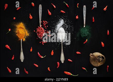 Verschiedene Arten von Gewürzen auf schwarzem Hintergrund in Teelöffeln dargestellt. Draufsicht über Kurkuma, roten Pfeffer, Chili, Salz und Spirulina Pulverfarbe Stockfoto