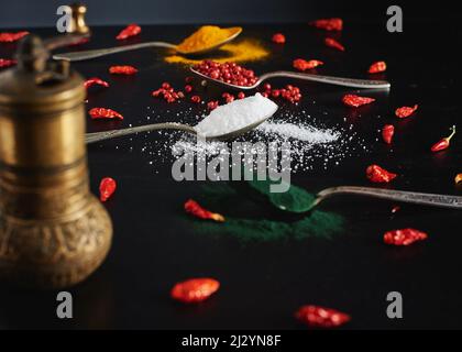 Roter Pfeffer, chilli, weißes Salz und Spirulina in Vintage-Löffeln auf schwarzem Hintergrund. Anordnung verschiedener Gewürzsorten in Tee sp Stockfoto