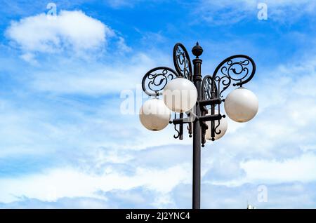 Straßenlampe auf dem Hintergrund des Himmels. Weiße kugelförmige Lampen, Eisenmuster. Straßenbeleuchtung in der Stadt. Stockfoto