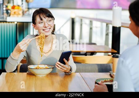 Porträt einer lächelnden jungen Frau, die köstliche asiatische Nudelgerichte im Café genießt und Wok mit Essstäbchen isst, Platz zum Kopieren Stockfoto