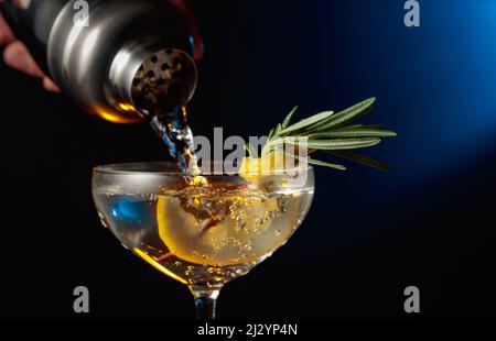 Gin Tonic mit Rosmarin und Zitrone. Der Cocktail wird aus einem Shaker in ein Glas gegossen. Stockfoto