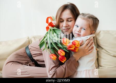 Freche kleine Mädchen wünscht ihrer Mutter alles gute zum Geburtstag, dem 8. März oder Muttertag, gibt Blumen, Küsse und Umarmungen Stockfoto