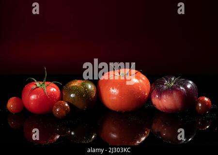 Bunte Tomaten in verschiedenen Größen und Arten auf rot dunklem Hintergrund mit Kopierraum. Stockfoto