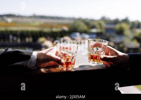Nahaufnahme von zwei Händen von Männern in Anzügen mit Gläsern Whiskey-Drink im Freien. Alkoholisches Getränk. Hochwertige Fotos. Stockfoto