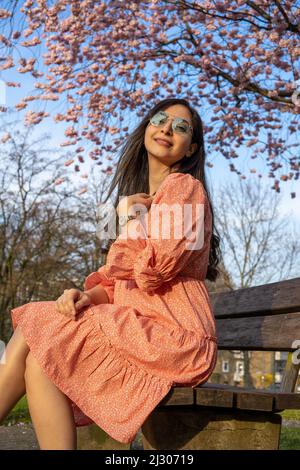 Schöne iranische junge Frau mit Sonnenbrille und buntem Kleid im Frühjahr mit einem Kirschblütenbaum auf dem Hintergrund Stockfoto