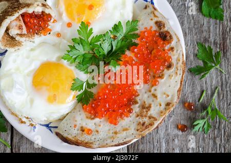 Spiegeleier mit Pfannkuchen und rotem Kaviar, Nahaufnahme, auf einem alten Holzhintergrund. Blick von oben Stockfoto