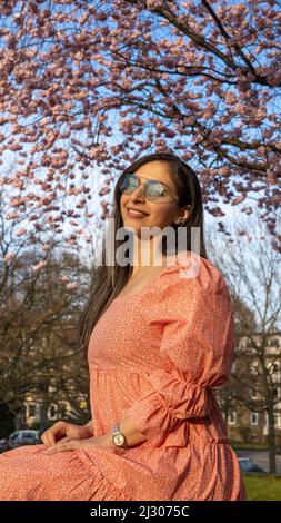 Schöne iranische junge Frau mit Sonnenbrille und buntem Kleid im Frühjahr mit einem Kirschblütenbaum auf dem Hintergrund Stockfoto