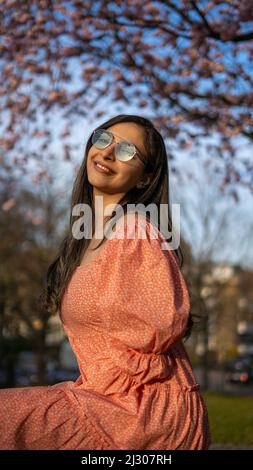 Schöne iranische junge Frau mit Sonnenbrille und buntem Kleid im Frühjahr mit einem Kirschblütenbaum auf dem Hintergrund Stockfoto