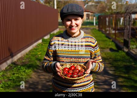 Unschärfe ukrainische junge lächelnde Frau in Hut hält Platte der Sammlung von rotem Ei auf Natur Hintergrund. Ostern, Ukraine. Bemalte Eier basteln. Pysanka Stockfoto