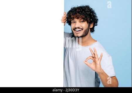 Glücklicher Inder oder Araber, in einem lässigen T-Shirt, der hinter dem Whiteboard der Werbung herauspiegt, einen leeren Platz für Text oder Design vorführt, zeigt eine GUTE Geste auf einem isolierten blauen Hintergrund Stockfoto