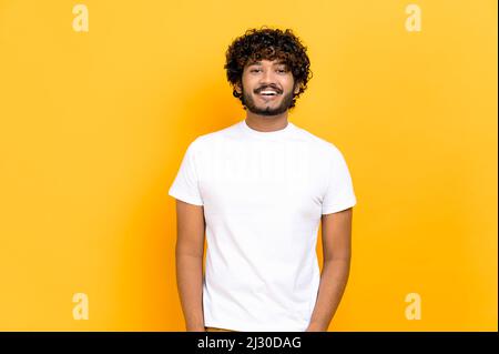 Porträt eines attraktiven, positiv gelockten indianers oder arabers, mit weißem Basic-T-Shirt, über einem isolierten orangefarbenen Hintergrund stehend, mit einem freundlichen Lächeln auf die Kamera schauend Stockfoto