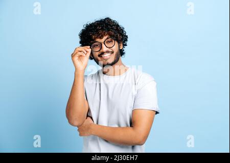 Attraktiver gutaussehender, positiver indischer oder arabischer trendiger lockiger Kerl mit Brille, Freiberufler oder kreativer Designer, steht auf blauem, isoliertem Hintergrund, blickt auf die Kamera, lächelt freundlich Stockfoto