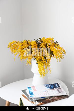 Vase mit Mimosenblumen und Zeitschriften auf Nachttisch in der Nähe einer hellen Wand Stockfoto