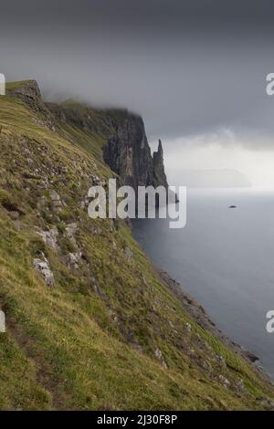 Blick entlang der Klippen zu Witches Finger, Meer Stapel. Vagar, Färöer. Dunkle Regenwolken. Stockfoto